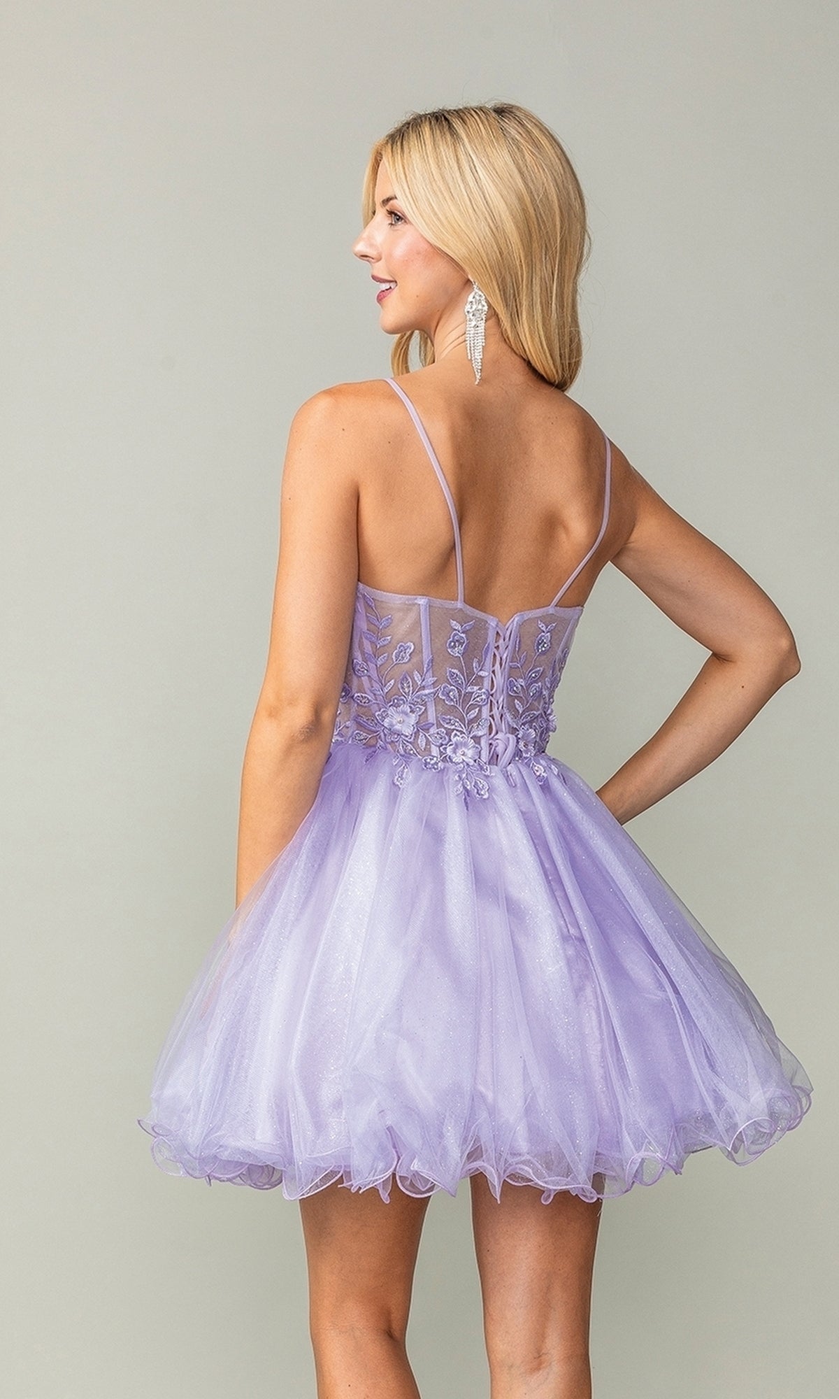 A woman with long blonde hair is shown from the back wearing the Sheer-Waist Short Babydoll Homecoming Dress 3382: a short, light purple tulle dress with thin straps and floral embroidery.
